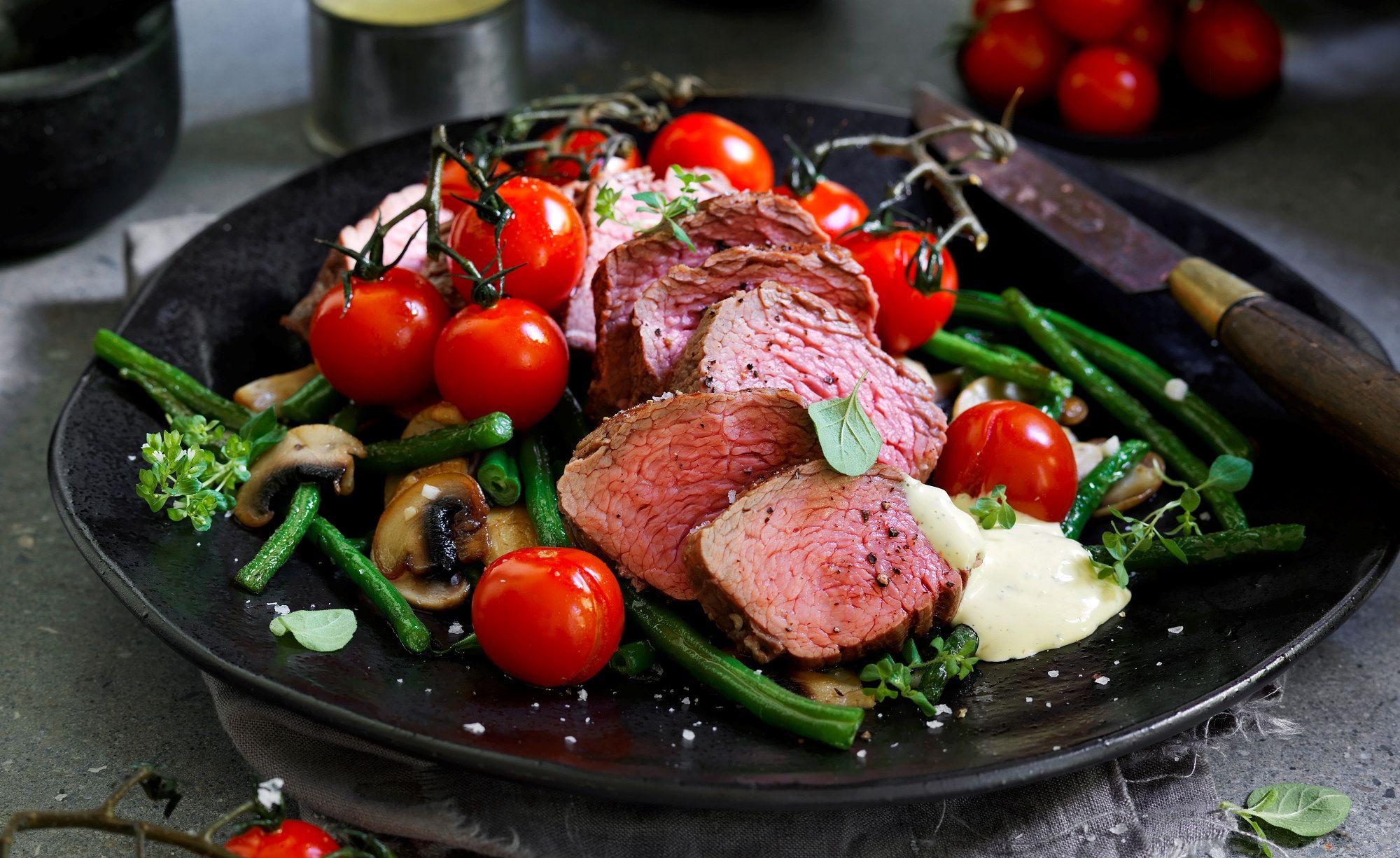 Stekt kjøtt med grønnsaker og cherrytomater på en svart tallerken.
