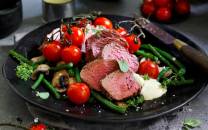 Stekt kjøtt med grønnsaker og cherrytomater på en svart tallerken.