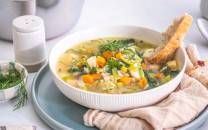 En bolle med grønnsaksuppe med gulrøtter, selleri og grønne urter, omgitt av brød og krydderurter på et bord.