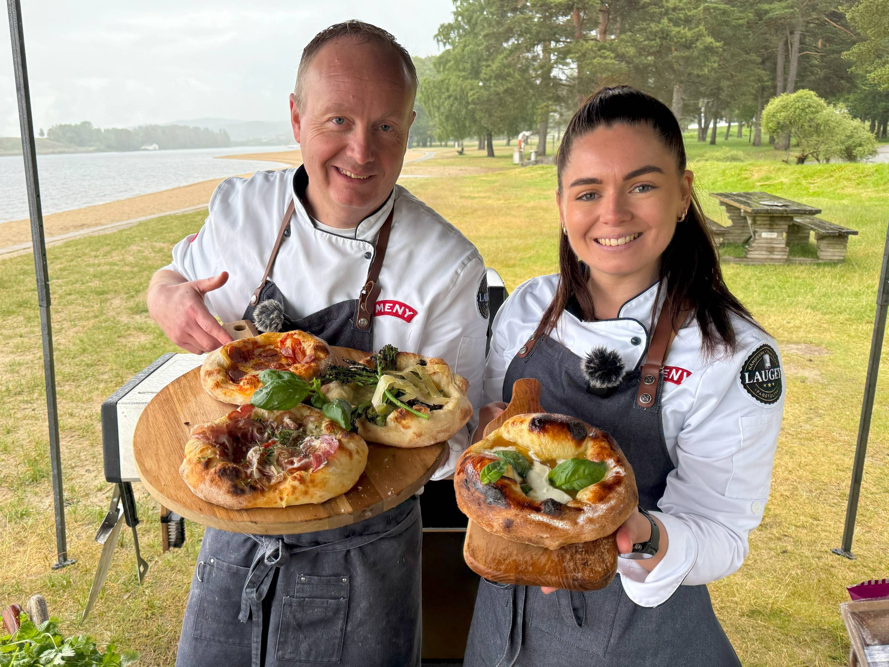 To personer som holder opp hver sin pizza med grønnsaker og kjøtt, utendørs ved en park eller innsjø, med trær og benker i bakgrunnen.