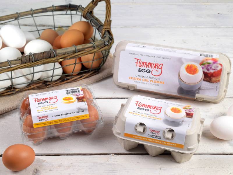 Egg i ulike farger og emballasje på et hvitt bord, med en kurv full av egg i bakgrunnen.