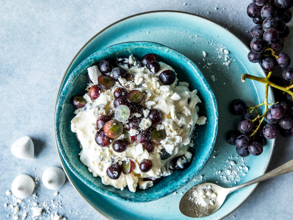 Pikekysskrem med druer i en blå bolle, omgitt av drueskudd og en skje med melis på et lyst bord.
