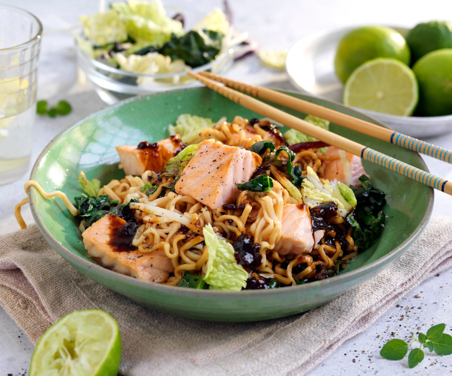 Wok med laks, nudler og grønnsaker i en grønn bolle, med lime og sitron i bakgrunnen