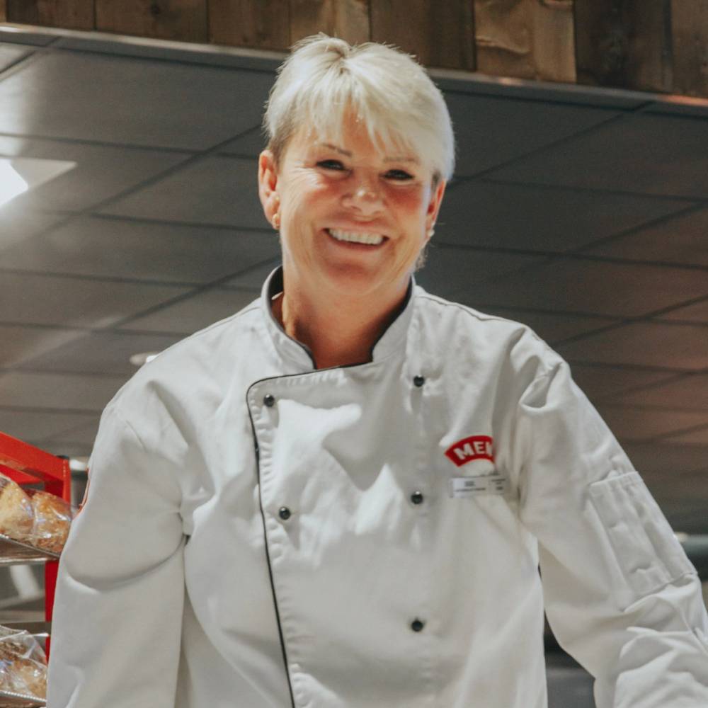 En person i hvit kokkeuniform står i et kjøkkenmiljø med tre hyller og matvarer i bakgrunnen.