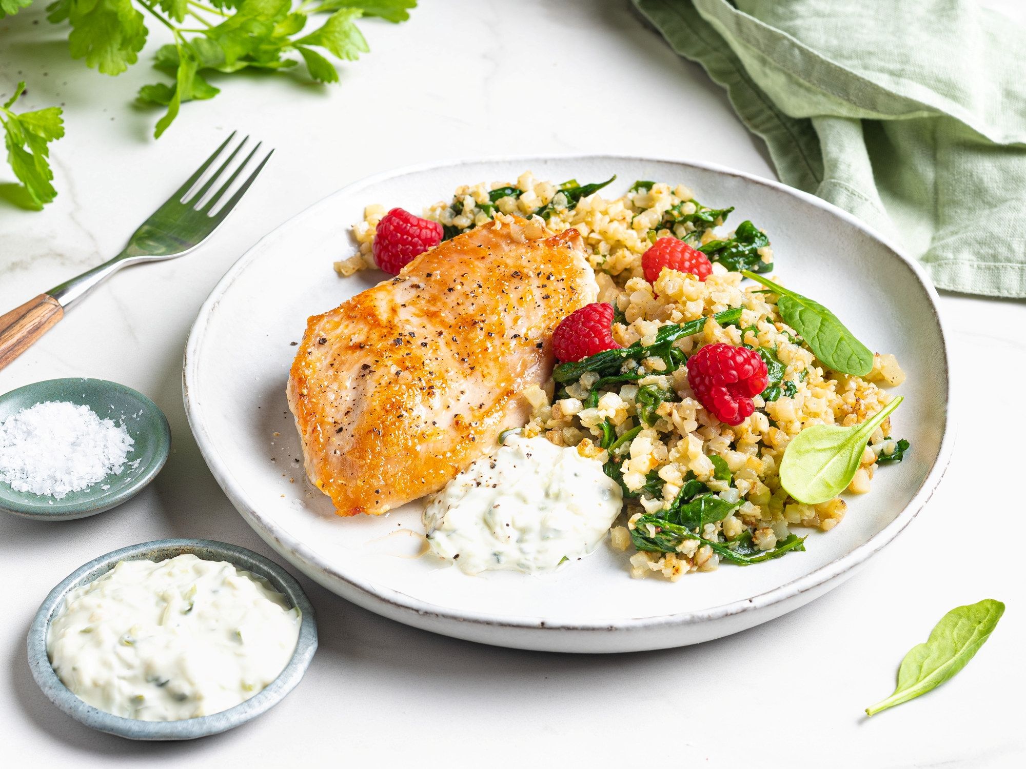 Kyllingfilet med blomkålris og friske bringebær på en hvit tallerken, omgitt av små boller med saus og en gaffel.