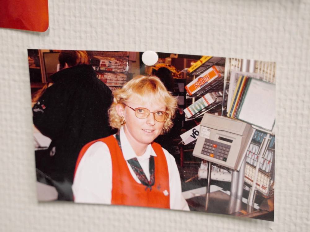 En ung person med lyst hår og rød vest står i en butikk, sannsynligvis i en matbutikk, med en kasseapparat og hyller med varer i bakgrunnen.