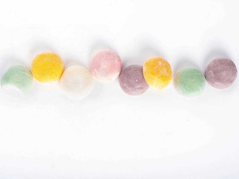 Fargede små sukkerkuler på rekke, med ulike farger som grønn, gul, rosa, hvit, lilla og brun, mot en hvit bakgrunn.