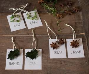 Navnelapper med navnene Lise, Bård, Julia, Lasse, Hanne og Erik, dekorert med grønne blader og stjerneanis, lagt på en treoverflate.