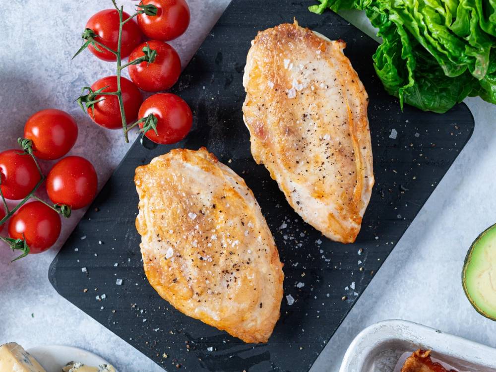 To stekte kyllingbryst med salt og pepper på en svart skjærefjøl, omgitt av cherrytomater, salat og avokado.