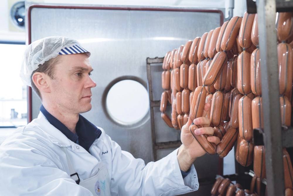 En person i hvit arbeidsdress og hårnet som sjekker pølser på en produksjonslinje i en fabrikk, med mange pølser på stativ i bakgrunnen.