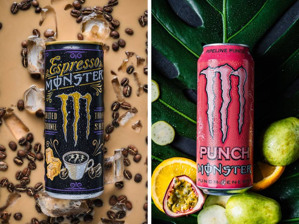 To energidrikker med Monster-logo, en med sjokoladesmak og en med punchsmak, omgitt av frukt og snacks.