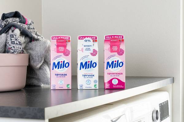 Tre pakker med Milo vaskemiddel står oppstilt på en kjøkkenbenk ved siden av en vaskemaskin, med en kurv med klær til venstre.