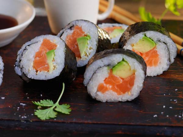 Fire maki-ruller med laks og avokado på en treplate, omgitt av soyasaus og grønne urter