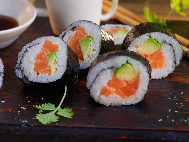 Fire maki-ruller med laks og avokado på en treplate, omgitt av soyasaus og grønne urter