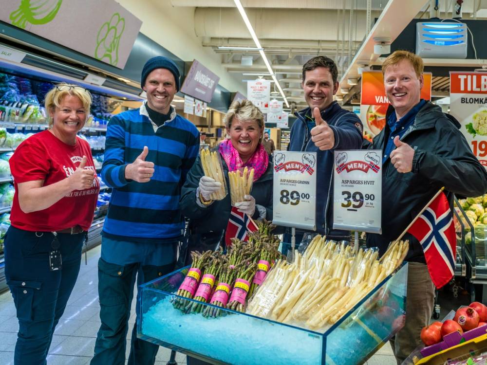 Gruppemennesker med blikkfang i en dagligvarebutikk, deriblant en person med norsk flagg, som står rundt en frukt- og grønnsaksdisk.