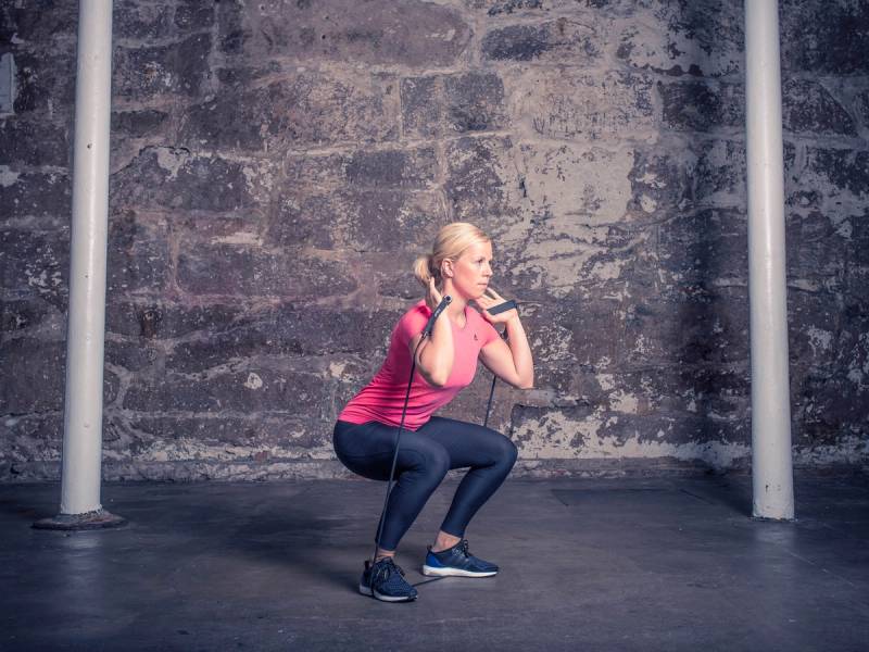 En person i rosa treningtøy gjør en knebøy i et treningsrom med en murvegg i bakgrunnen.