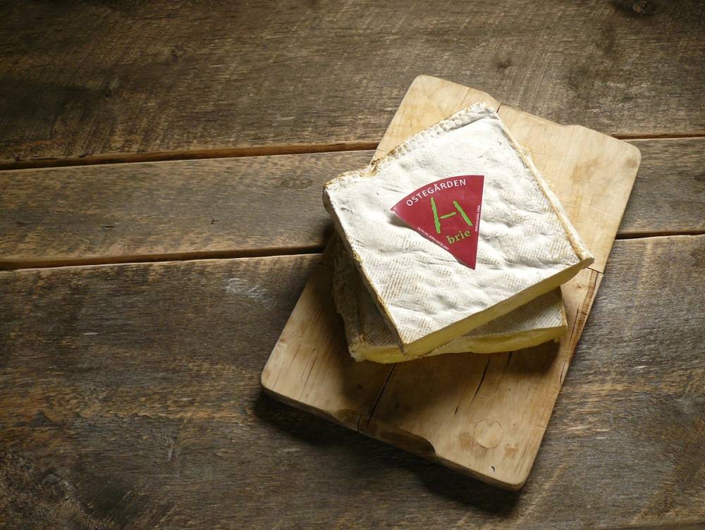 Et stykke ost med en rød og grønn etikett ligger på en skjærefjøl av tre på et trebord.