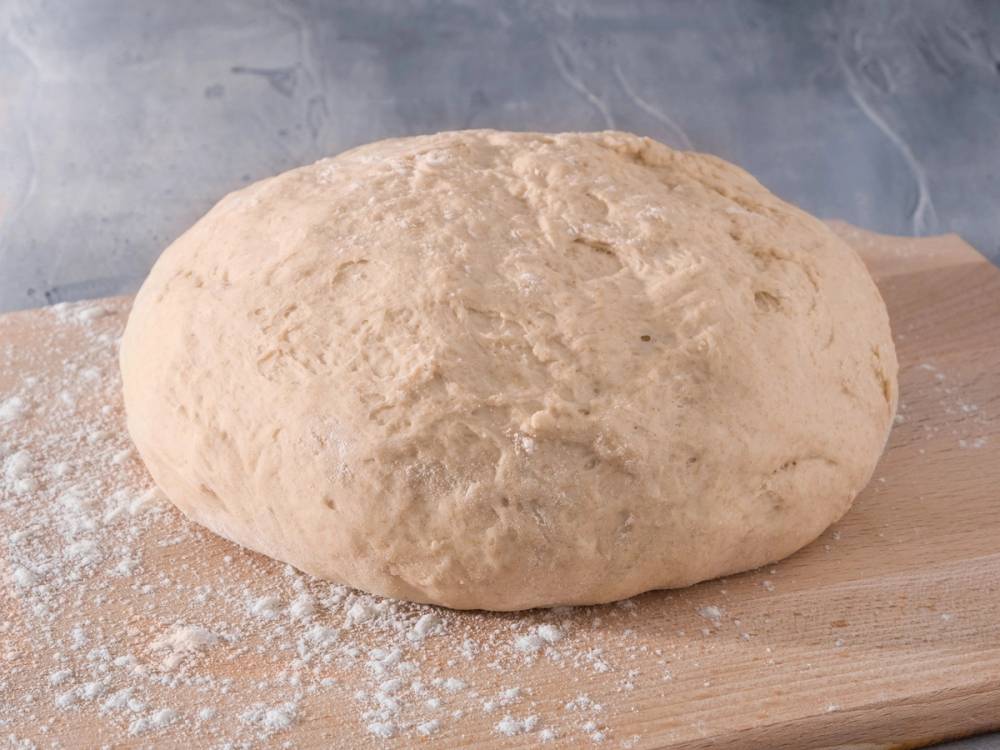En rund gjærdeig som er klar til å bli brukt til pizza, plassert på en treplate med litt mel rundt.