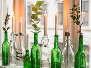 Flere grønne og klare glassflasker med lysende lysstaker på en vindusrekke, pyntet med grønne blader og små røde bær, som skaper en julestemning.