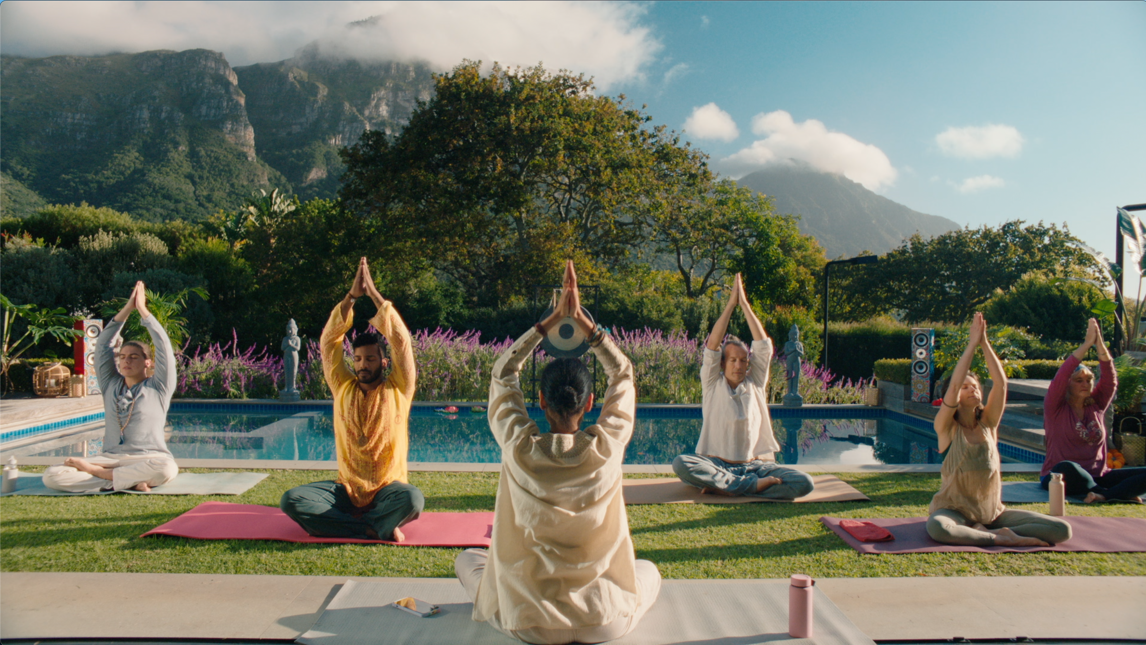 Fem personer som deltar i en utendørs yogaøkt på en plattform med fjell og trær i bakgrunnen, alle i en meditasjonsposisjon med hendene over hodet.
