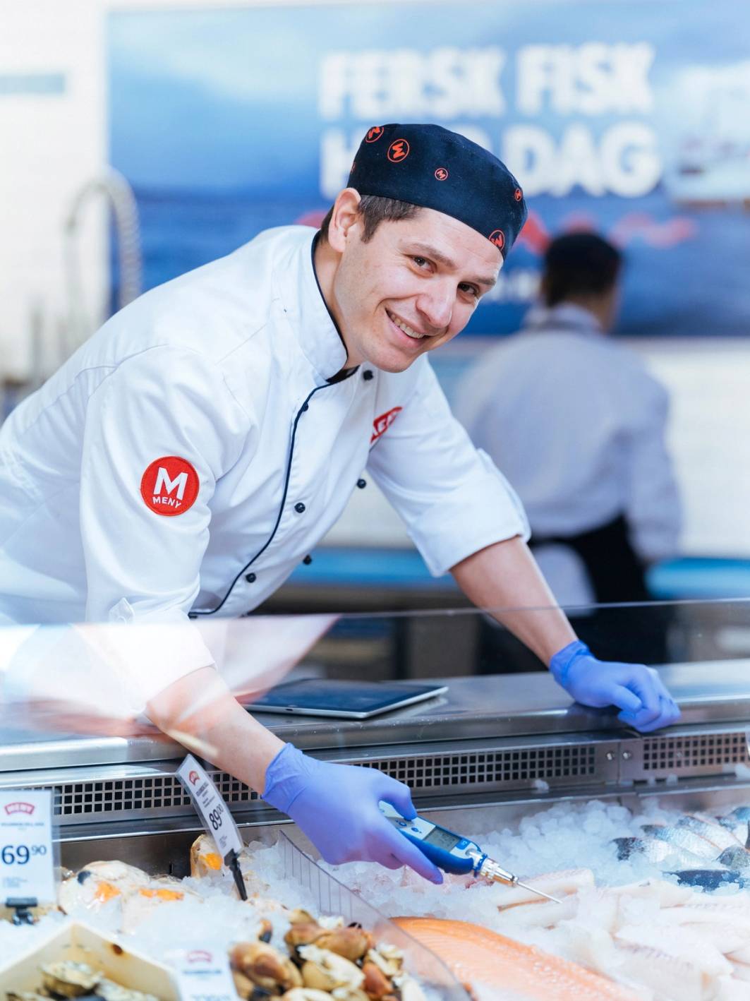 En kok i hvit uniform og blå hansker skjærer opp fisk på en fiskedisk, med en blå bakgrunn og tekst om fiskedag.