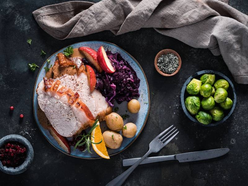 Skinkestek med tilbehør på tallerken, omgitt av grønnsaker og en liten skål med brune kuler