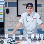 En person i kokkeforkle står bak en fiskedisk i en matbutikk, med ulike fisker og priser på displayet foran seg.