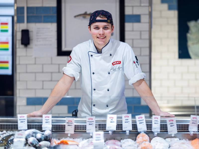 En person i kokkeforkle står bak en fiskedisk i en matbutikk, med ulike fisker og priser på displayet foran seg.