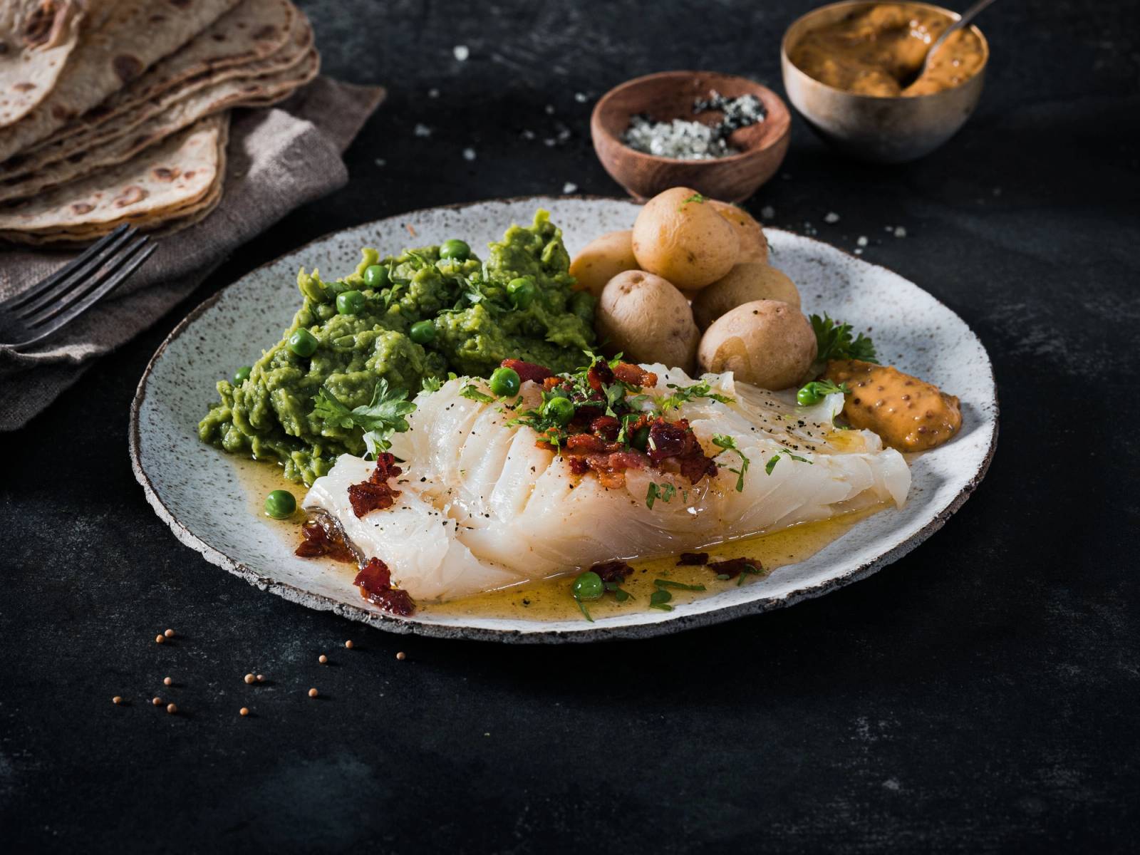 Lutefisk servert med grønnsaker og poteter på en hvit tallerken, med små skåler med tilbehør i bakgrunnen.