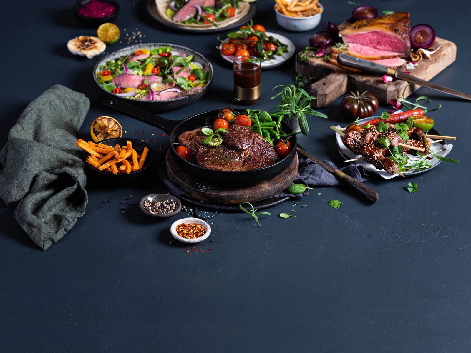 Et utvalg av ulike kjøttretter og tilbehør på en mørk bakgrunn, inkludert en biff og grønnsaker.