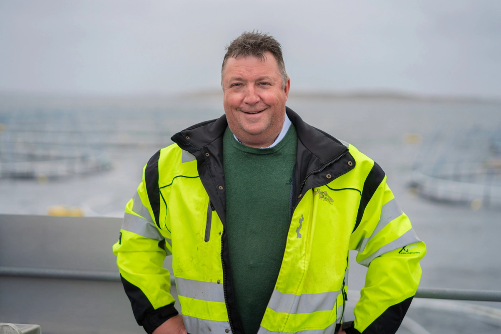 Person i gul refleksvest står ved sjøen med båter i bakgrunnen, utsikten er åpen og overskyet.