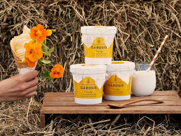 En person holder en blomsterbukett med oransje blomster foran en bord med tre bøtter med Gårdsis iskrem og en bolle med iskrem, mot en bakgrunn av tørt gress.