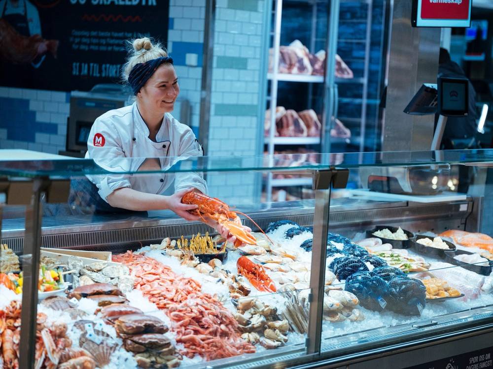 Kjøtt- og pølseutvalg i en kjøttbutikk med en ansatt som pakker kjøtt.