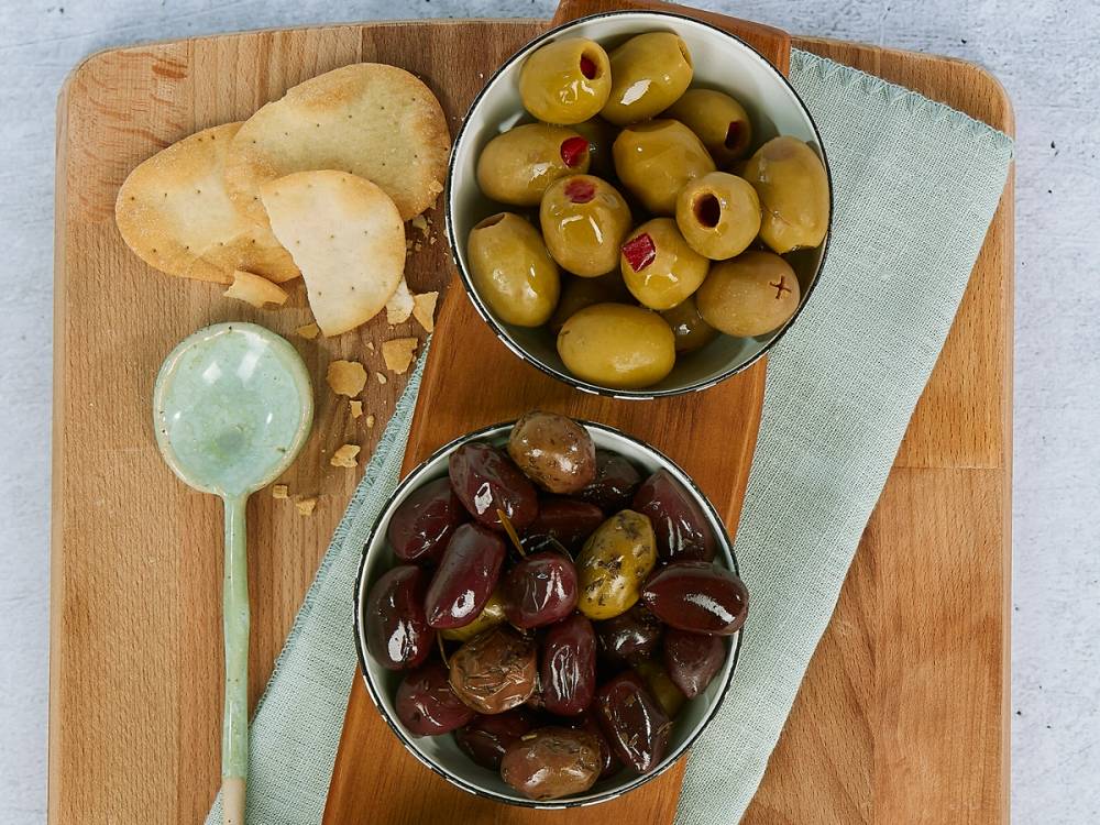 Skåler med grønne og svarte oliven på et trebord med kjeks og en skje ved siden av.
