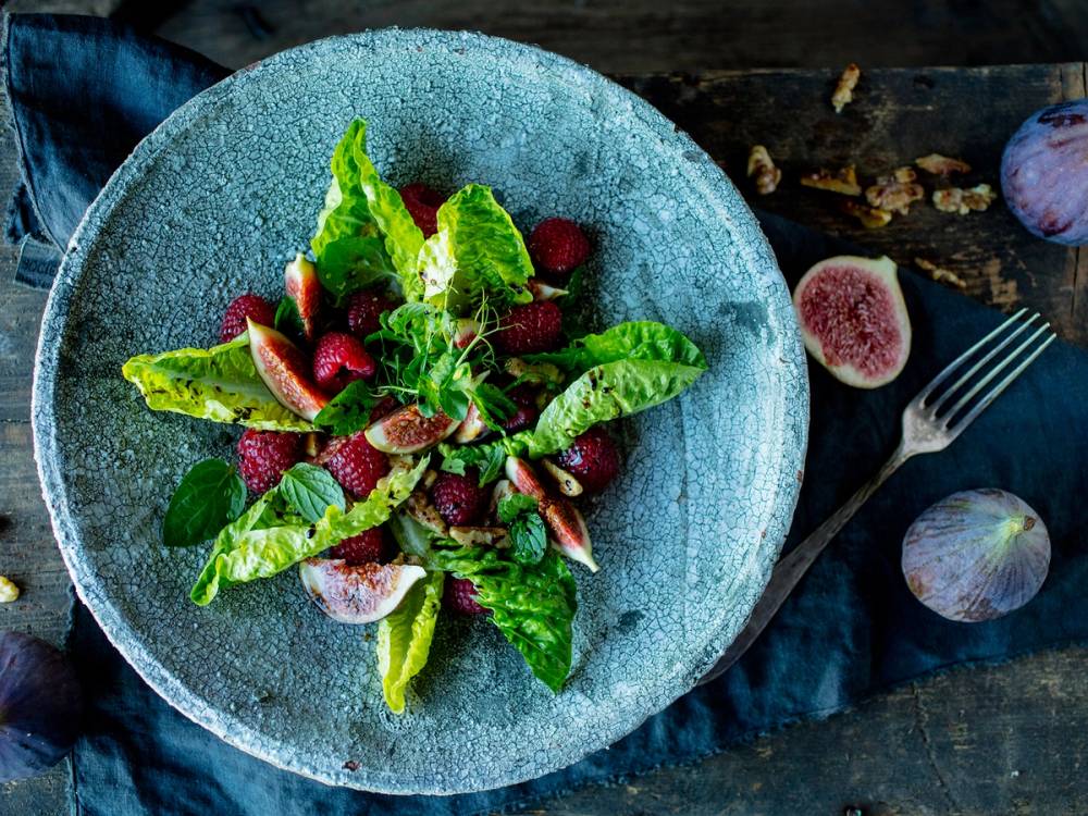 En salat med fiken, bringebær og grønne blader i en blå bolle på et mørkt bord.