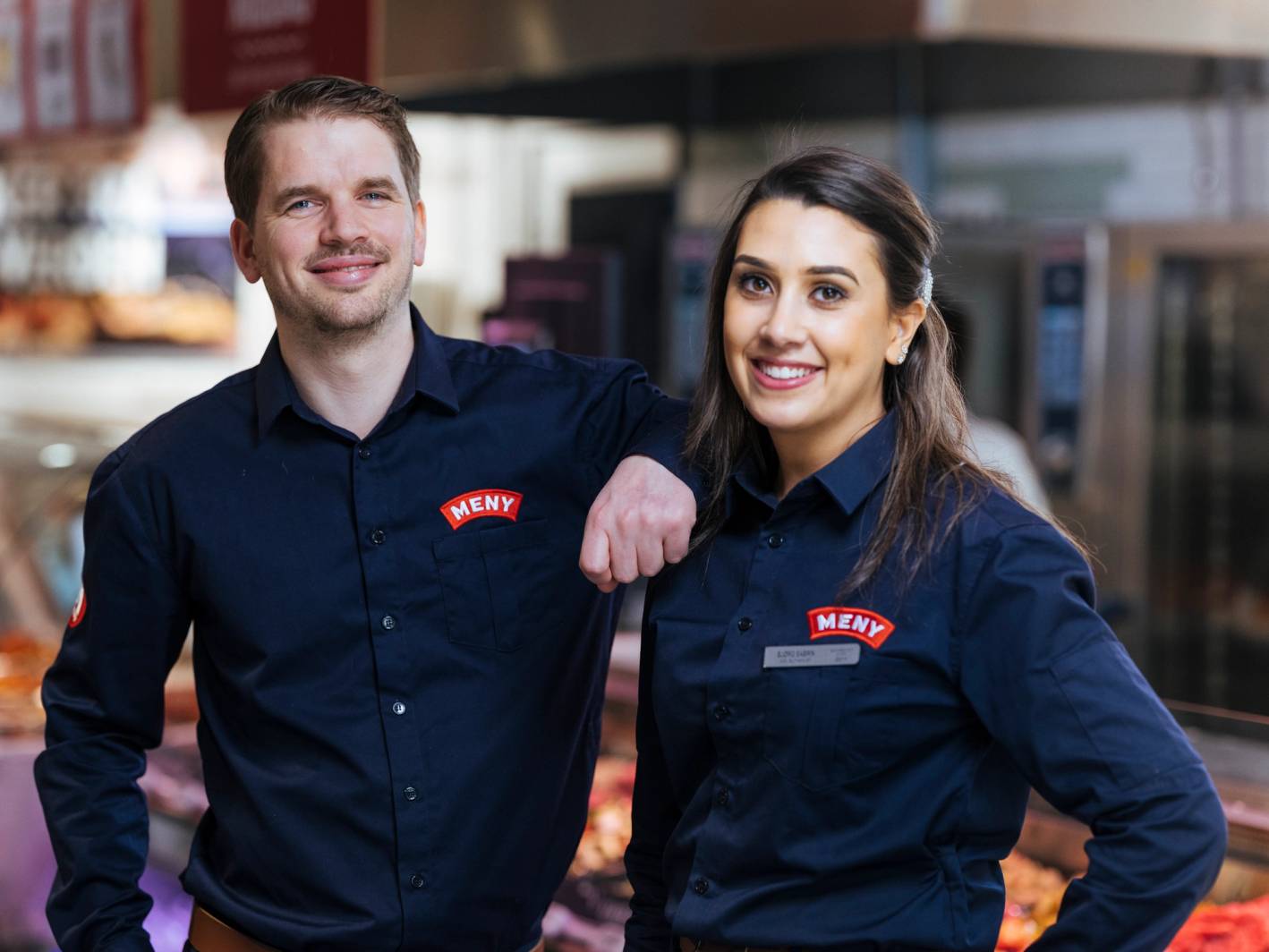 To ansatte i uniform med navnemerke står i en butikk eller restaurant, og den ene har armen rundt den andre.