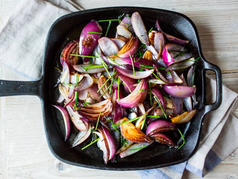 Skjult bilde av rødløk, hvitløk og timian i en svart stekepanne på et trebord.