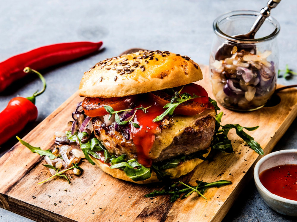 Hamburger med chili, ost og grønnsaker på et skjærebrett med chili og saus ved siden av.