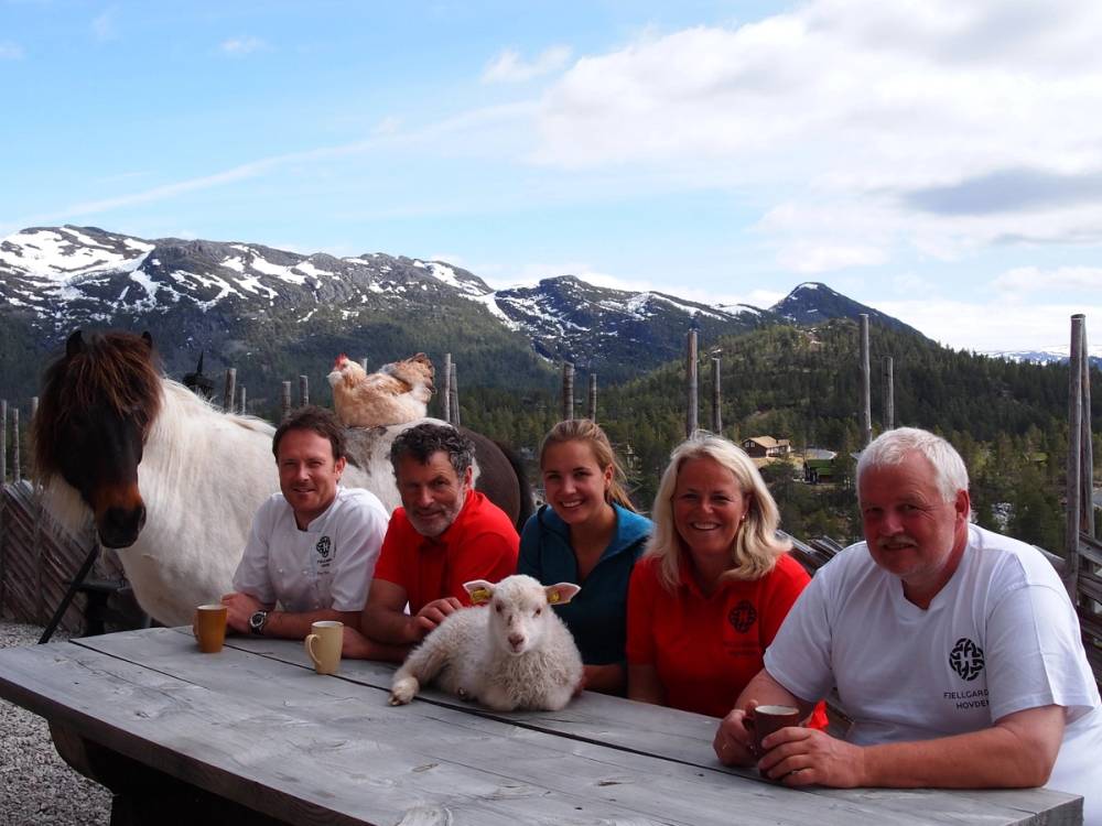 En gruppe mennesker og en hund sitter ved et bord med fjell og snø på toppen i bakgrunnen, på en klar dag.
