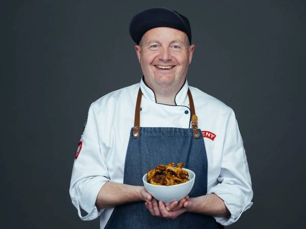 En person i kokkeuniform og forkle holder en bolle med mat, mot en mørk bakgrunn.