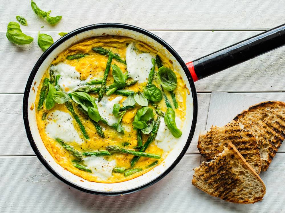 Omelett med asparges og egg i en stekepanne, med brød ved siden av, på et hvitt bord.