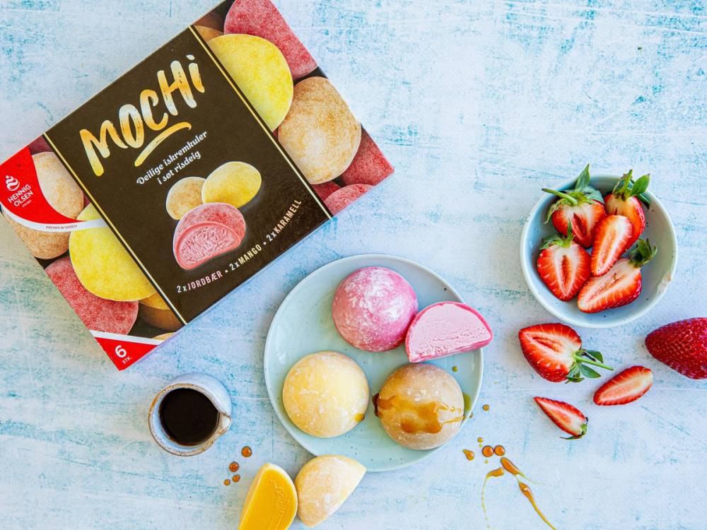 En plate med ulike fargede mochi-is, en eske med mochi i pakke, og en liten kopp med soyasaus på et lyst bord.