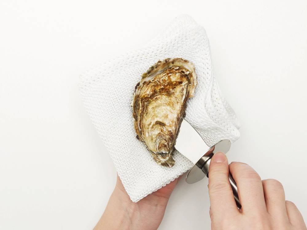 En person holder en østers med en liten kniv på en hvit klut, klar til å åpne den.
