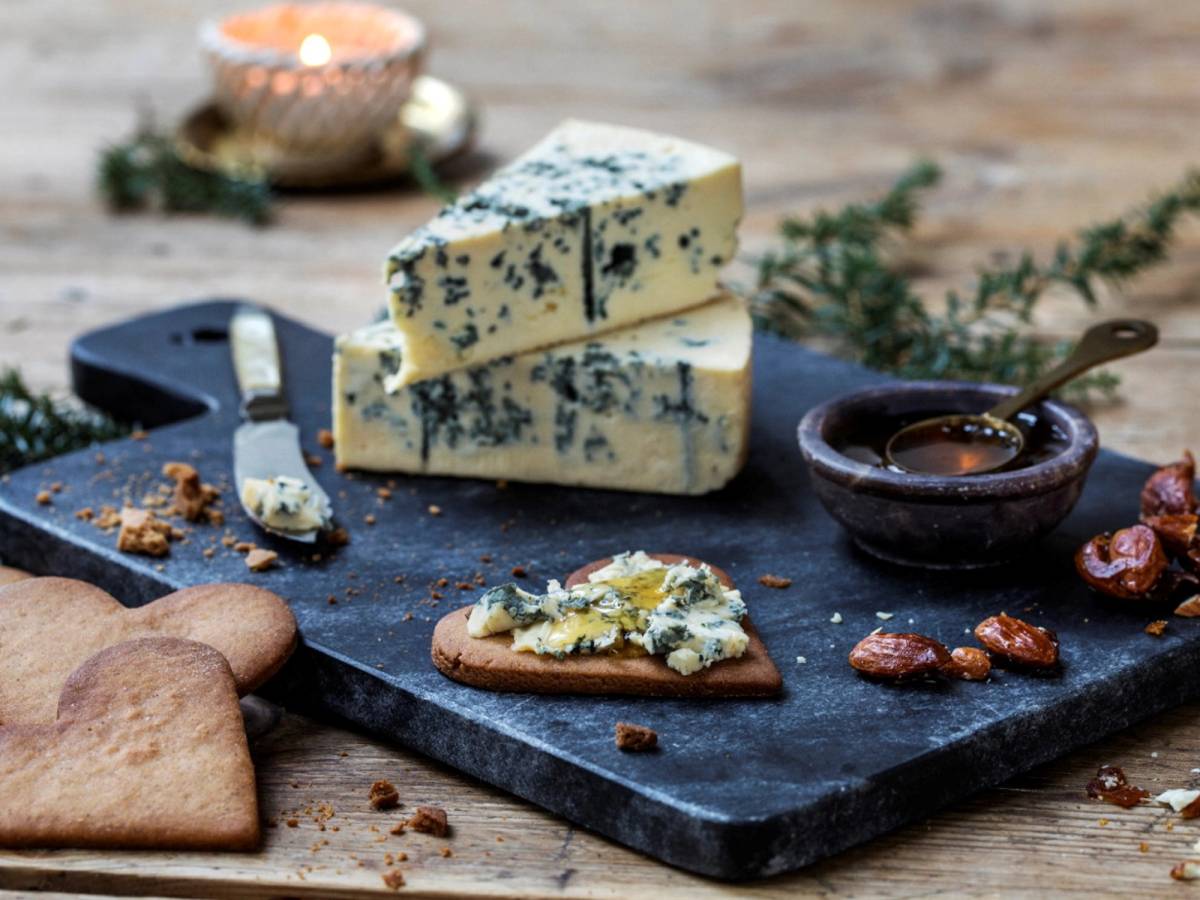 To stykker blåmuggost på en svart skjærefjøl med kjeks og honning, omgitt av nøtter og en liten lysestake i bakgrunnen.