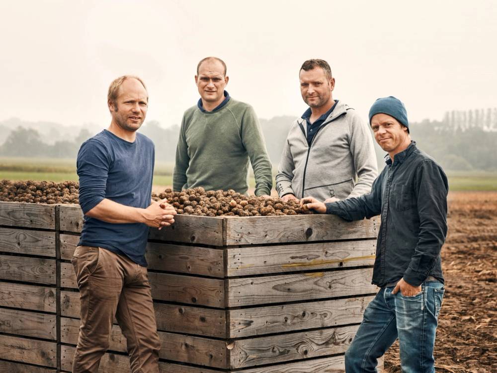 Fire personer står rundt en stor blomsterkasse fylt med jord i et utendørs landskap, og snakker sammen.