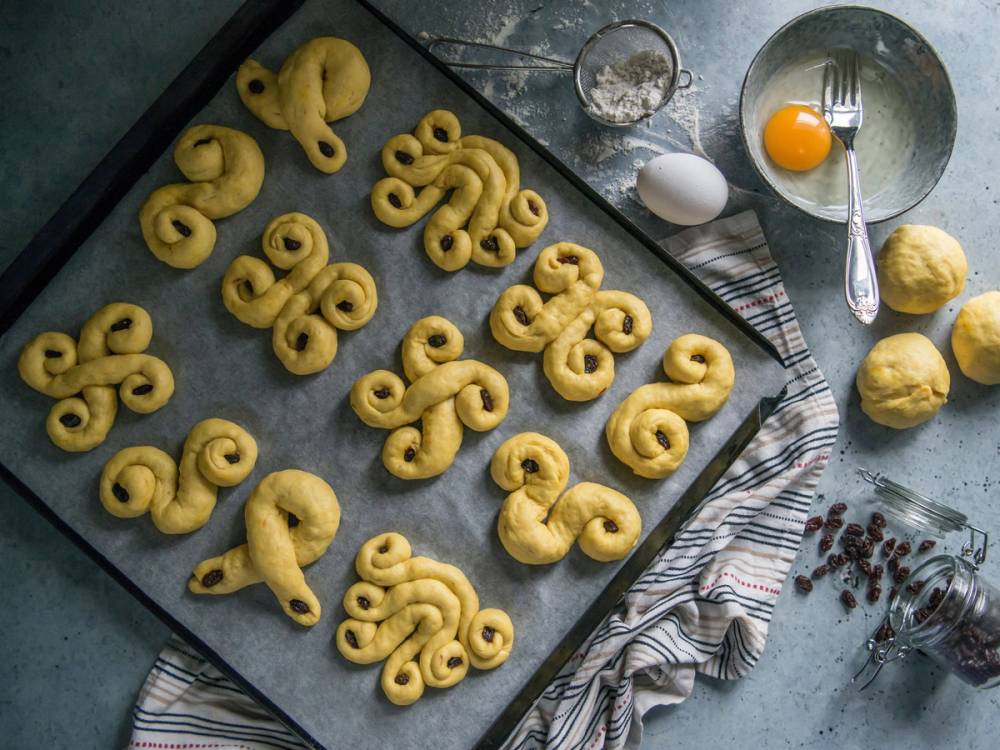 Lussekatteformede små bakverk på et stekebrett, klar til å stekes, med ingredienser og redskaper rundt.
