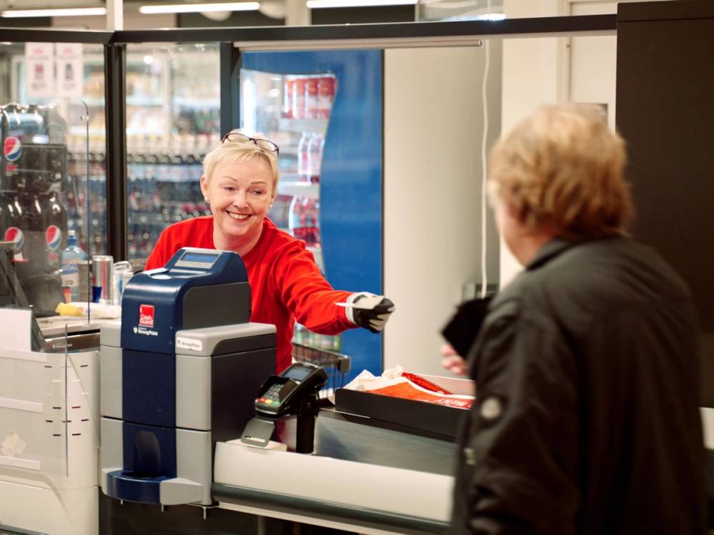 En kvinne i rød genser betjener en kunde i kassen på en dagligvarebutikk, med en annen kunde i bakgrunnen og butikkens utvalg av varer bak disken.