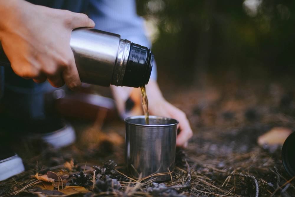 En person heller varm drikke fra en termos ned i et metallkrus utendørs i skogen.