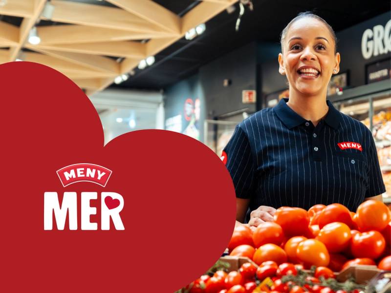En person står bak en fruktstasjon med tomater i en butikk, med MENY MER-logoen synlig i bakgrunnen.