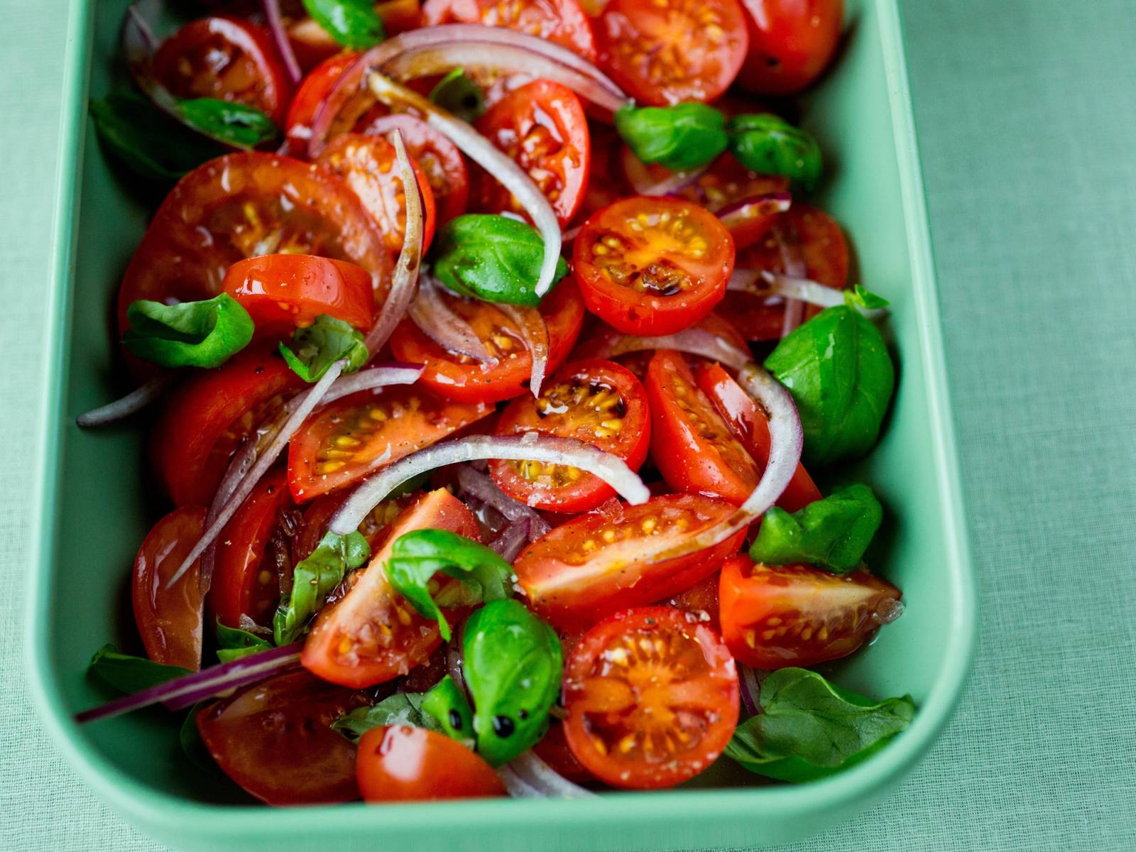 Cherrytomater delt i to, med noen ferske basilikumblader og skiver av rødløk i en grønn plastboks